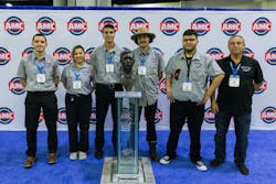 Five AMTs in grey shirts and one in a navy blue shirt stand smiling behind a bronze bust Five AMTs in grey shirts and one in a navy blue shirt stand smiling behind a bronze bust