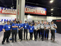 A group of technicians wearing blue and grey shirts pose in front of a banner that reads, 'Aerospace Maintenance Council COMPETITION' A group of technicians wearing blue and grey shirts pose in front of a banner that reads, 'Aerospace Maintenance Council COMPETITION'