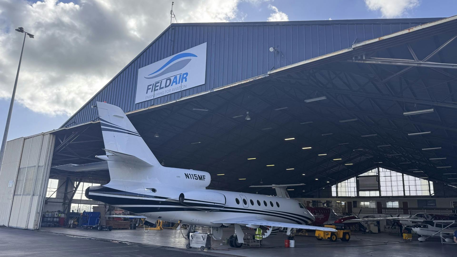 An aircraft hangar with the door open and an airplane parked halfway inside, with a sign on the front of the building that reads: FIELDAIR