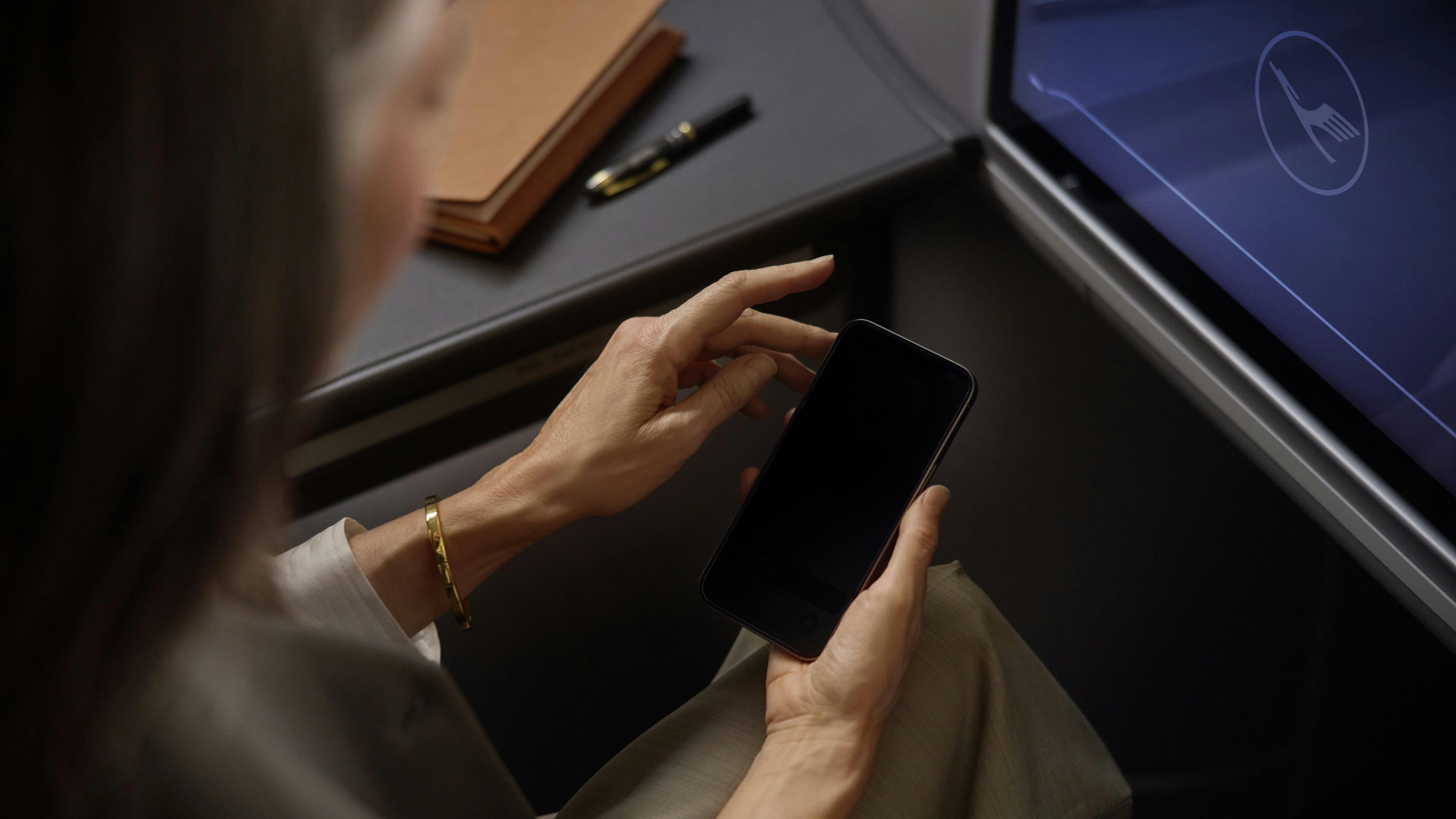 A woman holding a cell phone while sitting in an airplane seat with the Lufthansa logo on a screen on the back of the seat in front of her