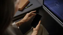 A woman holding a cell phone while sitting in an airplane seat with the Lufthansa logo on a screen on the back of the seat in front of her