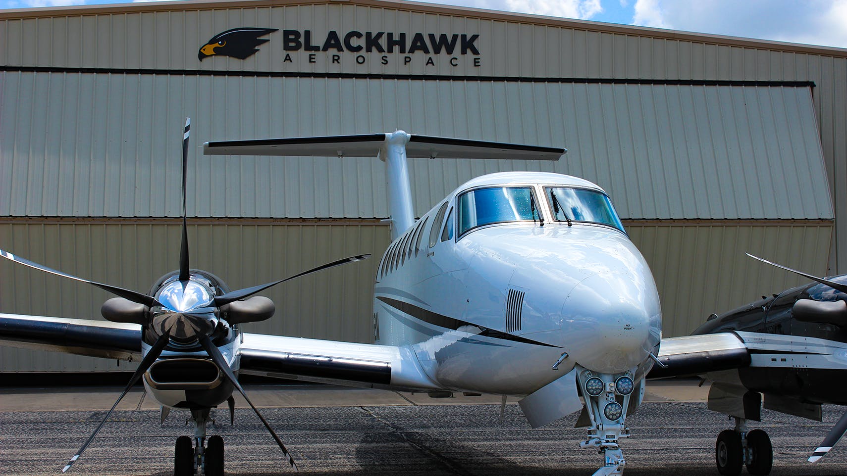 An airplane with a 5-blade propeller sits parked in front of an aircraft hangar with a sign on the front that reads: BLACKHAWK Aerospace