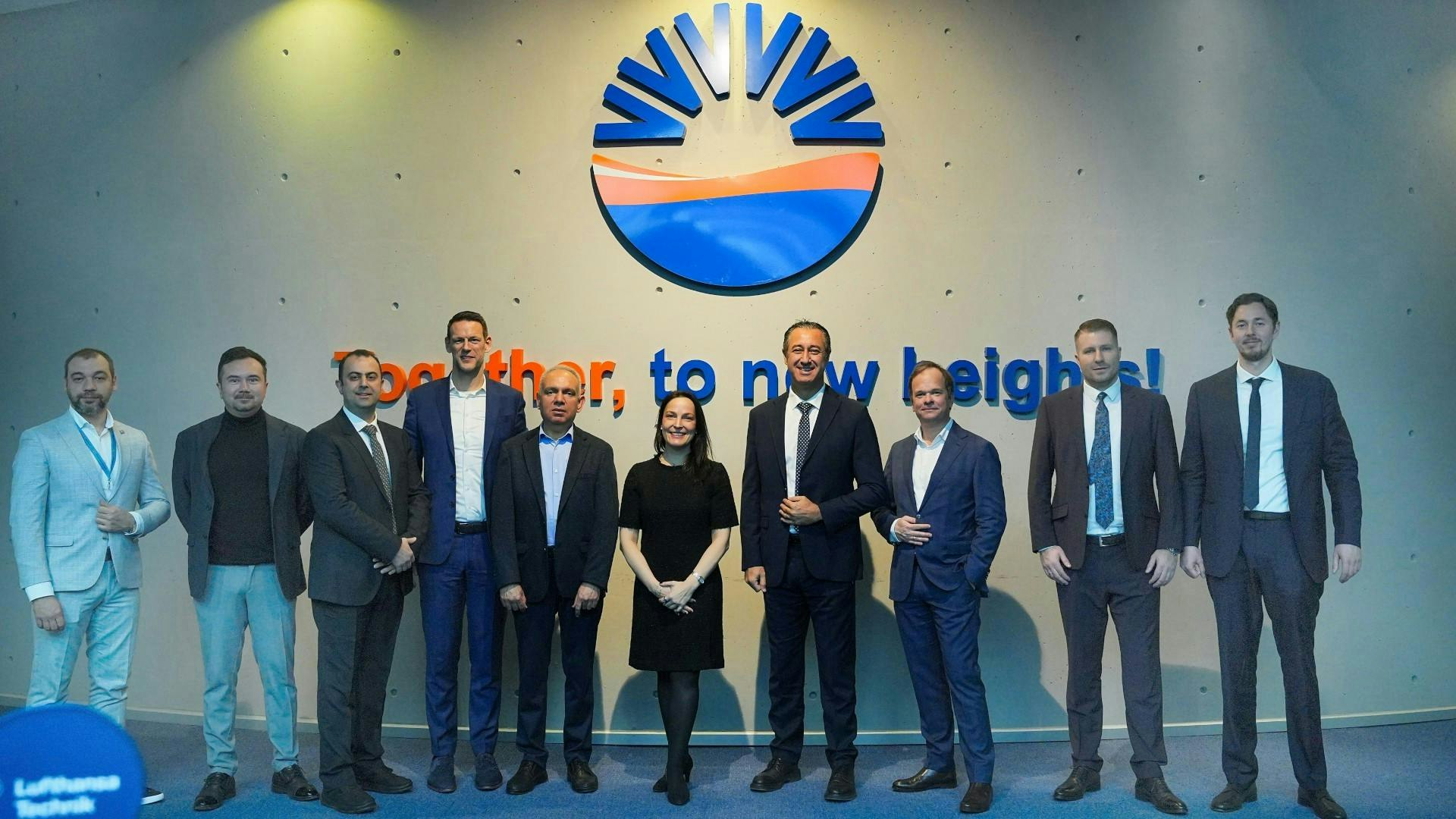 A group of professionals in business attire smiling at the camera and standing in front of a sign that reads: Together, to new heights