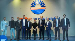 A group of professionals in business attire smiling at the camera and standing in front of a sign that reads: Together, to new heights