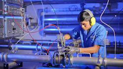 A maintenance technician wearing a blue shirt and yellow over-ear headphones using a piece of tech equipment to work on an airplane part A maintenance technician wearing a blue shirt and yellow over-ear headphones using a piece of tech equipment to work on an airplane part