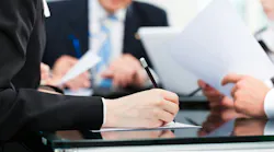 A close-up shot of a hand writing with a pen on a piece of paper, with other people in the background wearing suits, insinuating a meeting