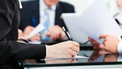 A close-up shot of a hand writing with a pen on a piece of paper, with other people in the background wearing suits, insinuating a meeting A close-up shot of a hand writing with a pen on a piece of paper, with other people in the background wearing suits, insinuating a meeting