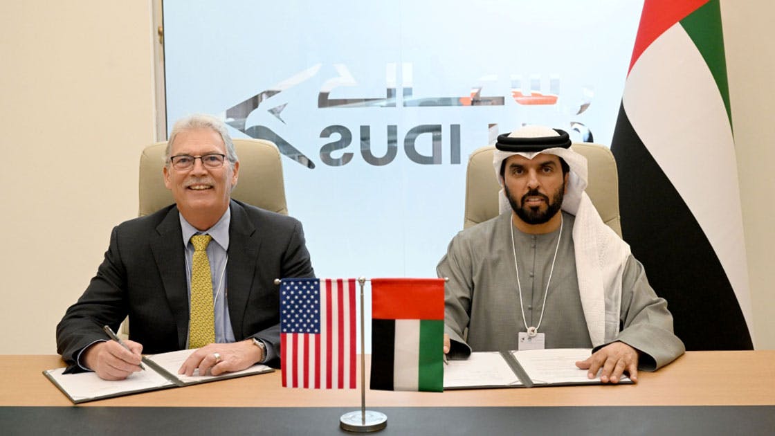 Two men in professional attire sitting at a desk while singing papers and smiling at the camera, with an American flag and a United Arab Emirates flag on the table in front of them