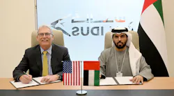 Two men in professional attire sitting at a desk while singing papers and smiling at the camera, with an American flag and a United Arab Emirates flag on the table in front of them