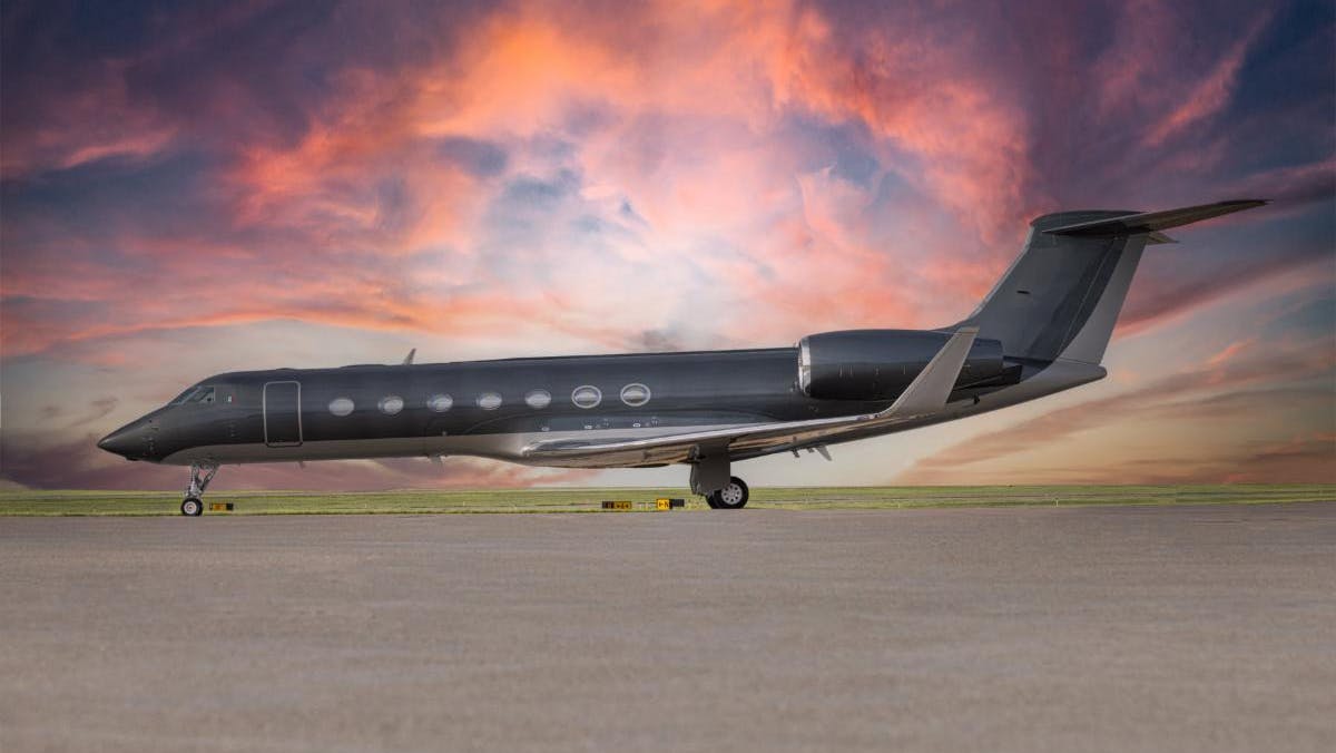 A dark gray airplane parked on a tarmac in front of a sky during sunset, with pink and purple hues in the clouds