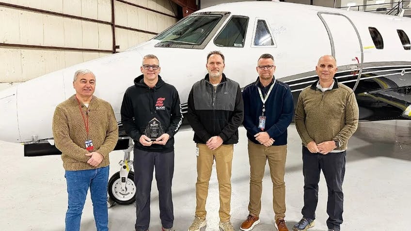 Four men wearing ID badges and casual clothing smiling at the camera while standing in front of an airplane, with one man holding an award