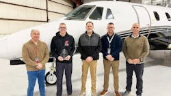Four men wearing ID badges and casual clothing smiling at the camera while standing in front of an airplane, with one man holding an award Four men wearing ID badges and casual clothing smiling at the camera while standing in front of an airplane, with one man holding an award