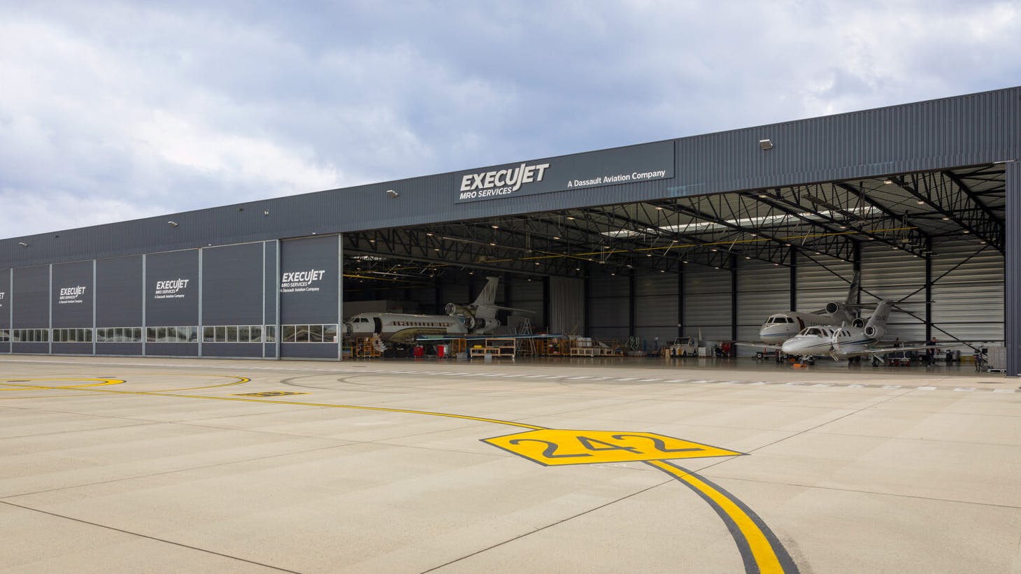 The outside of an aircraft maintenance hangar with the doors open and an airplane inside, with a sign on the front of the building that says: ExecuJet MRO Services