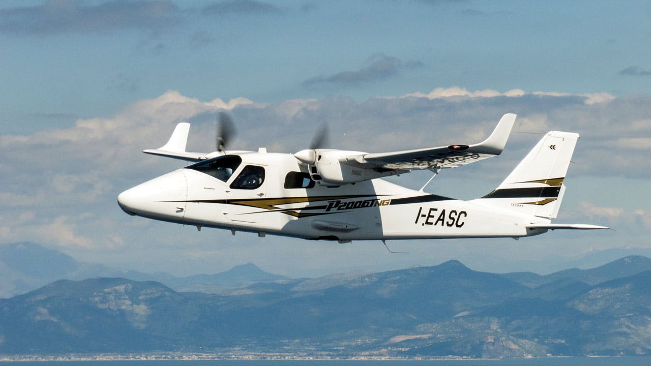 A white airplane with black and brown accents flying over green and brown mountains in front of a blue sky with white clouds