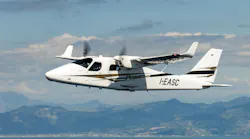 A white airplane with black and brown accents flying over green and brown mountains in front of a blue sky with white clouds