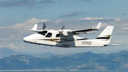 A white airplane with black and brown accents flying over green and brown mountains in front of a blue sky with white clouds A white airplane with black and brown accents flying over green and brown mountains in front of a blue sky with white clouds