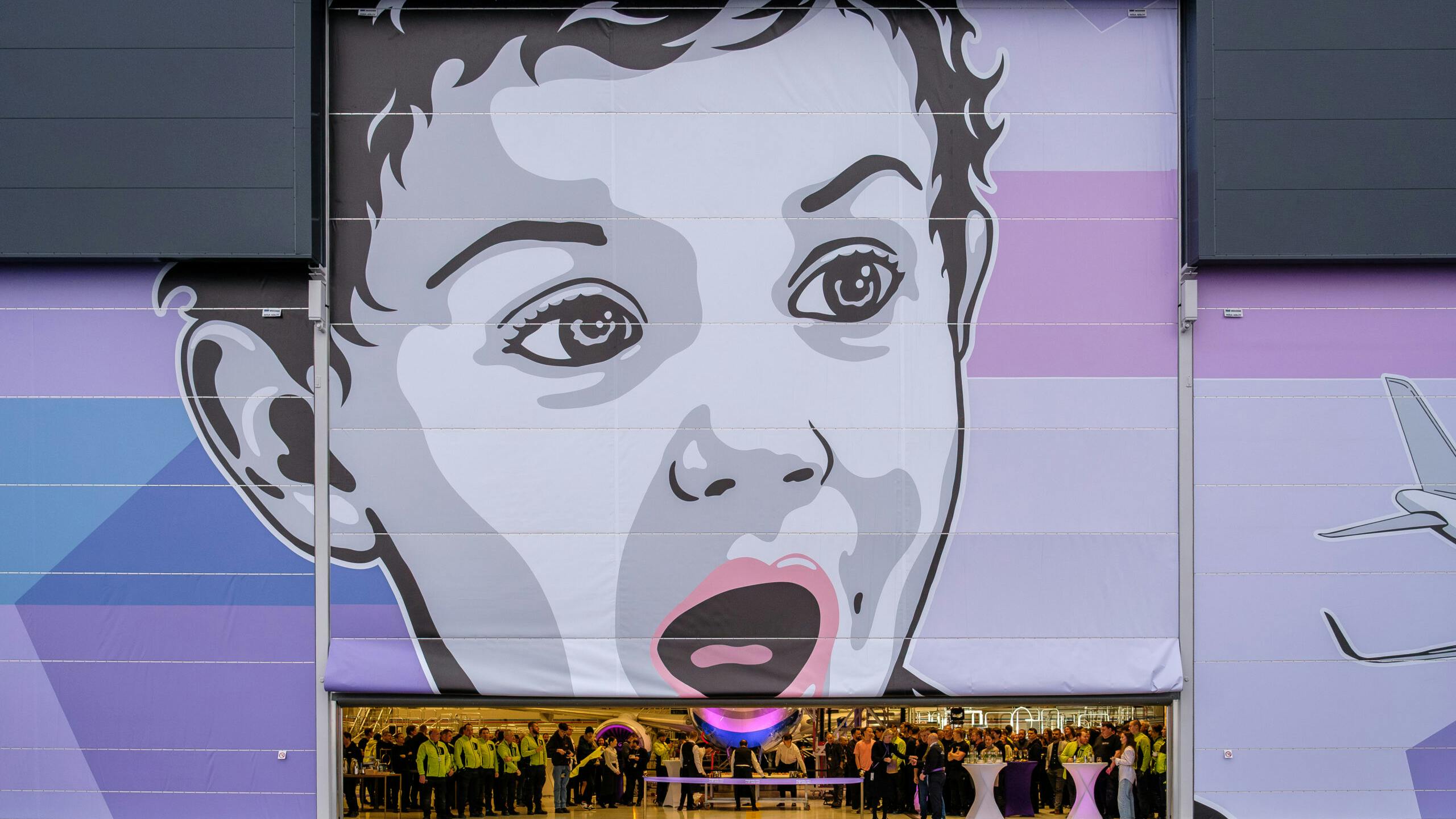 A large hangar door with a mural painted on it, showcasing a child's face and an airplane off to the side in shades of gray, blue and purple