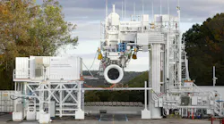 A large white piece of machinery testing an aircraft engine outdoors with autumnal trees next to it