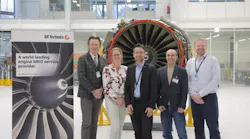 Five people wearing business attire and ID badges stand smiling in front of an airplane engine with a sign next to them that reads: SR Technics A world leading engine MRO service provider