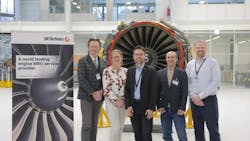 Five people wearing business attire and ID badges stand smiling in front of an airplane engine with a sign next to them that reads: SR Technics A world leading engine MRO service provider Five people wearing business attire and ID badges stand smiling in front of an airplane engine with a sign next to them that reads: SR Technics A world leading engine MRO service provider