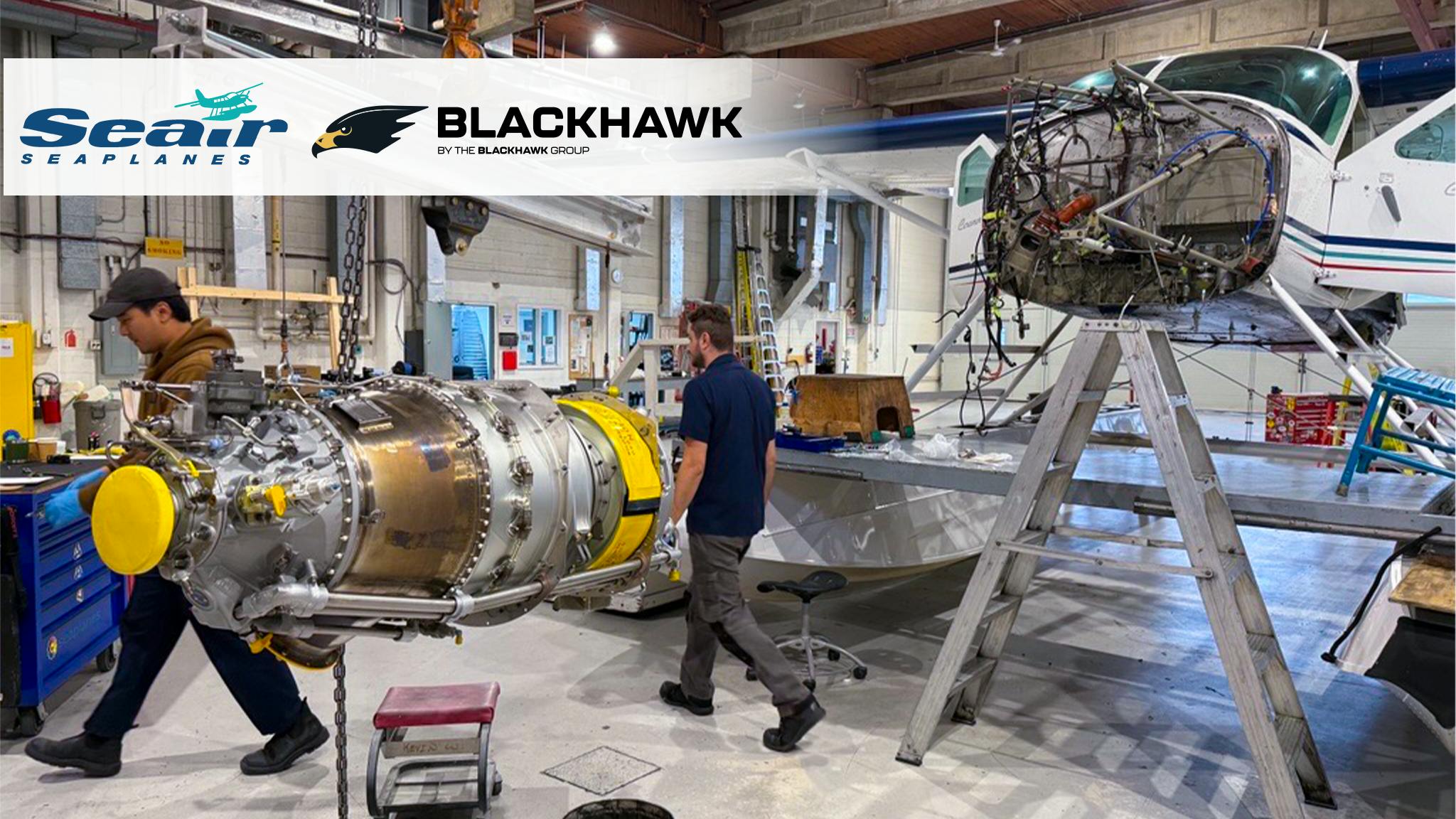 The interior of a maintenance hangar showing two technicians walking near an airplane that has been taken apart and an engine on a stand. There's text in the corner showing two logos that read: 'Seair Seaplanes' and 'Blackhawk by The Blackhawk Group'