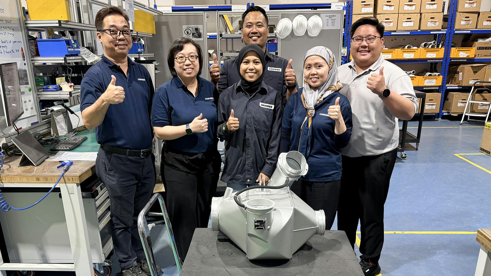 A group of maintenance technicians smiling at the camera with some giving a thumbs-up hand gesture, all standing behind an aircraft component in a repair shop
