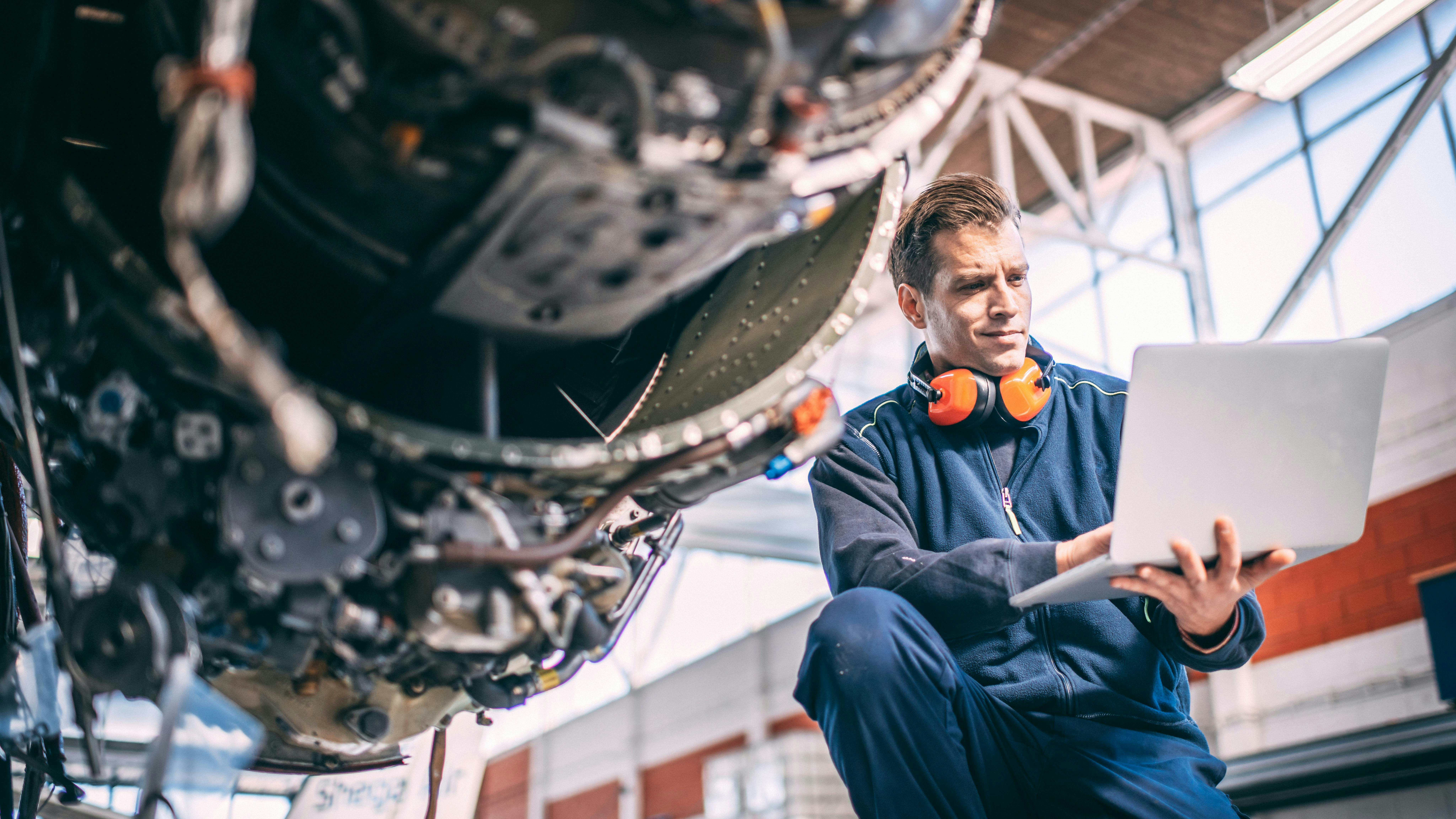 An aircraft maintenance technician wearing a blue uniform and orange over-ear headphones holds a computer and looks at it while crouching next to an exposed aircraft engine
