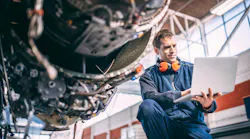 An aircraft maintenance technician wearing a blue uniform and orange over-ear headphones holds a computer and looks at it while crouching next to an exposed aircraft engine