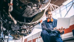 An aircraft maintenance technician wearing a blue uniform and orange over-ear headphones holds a computer and looks at it while crouching next to an exposed aircraft engine An aircraft maintenance technician wearing a blue uniform and orange over-ear headphones holds a computer and looks at it while crouching next to an exposed aircraft engine