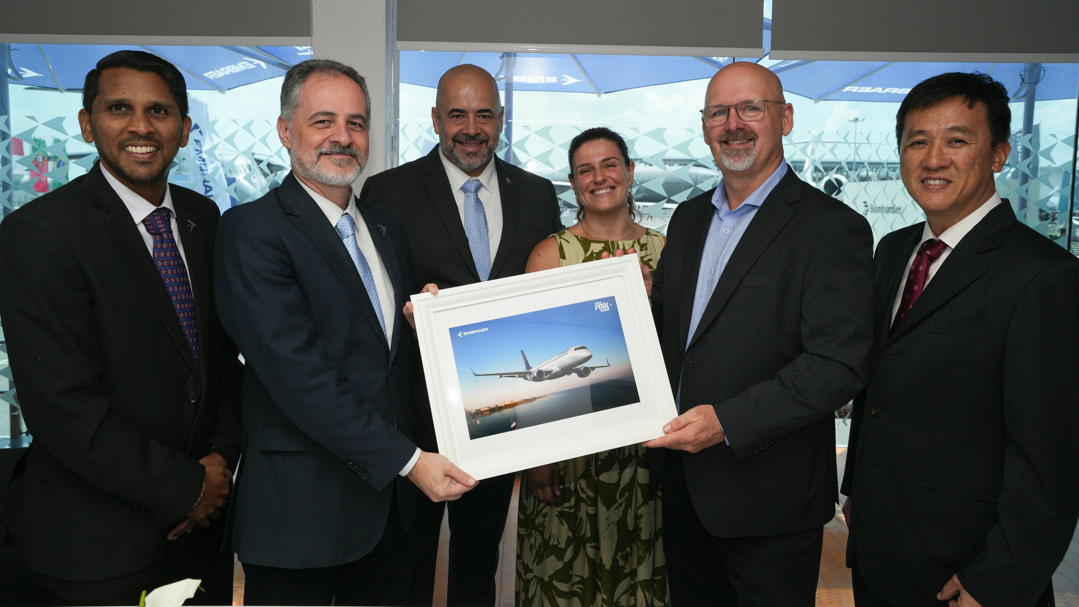 A group of professionals in business attire smile at the camera together, with two men holding a framed photo of an airplane