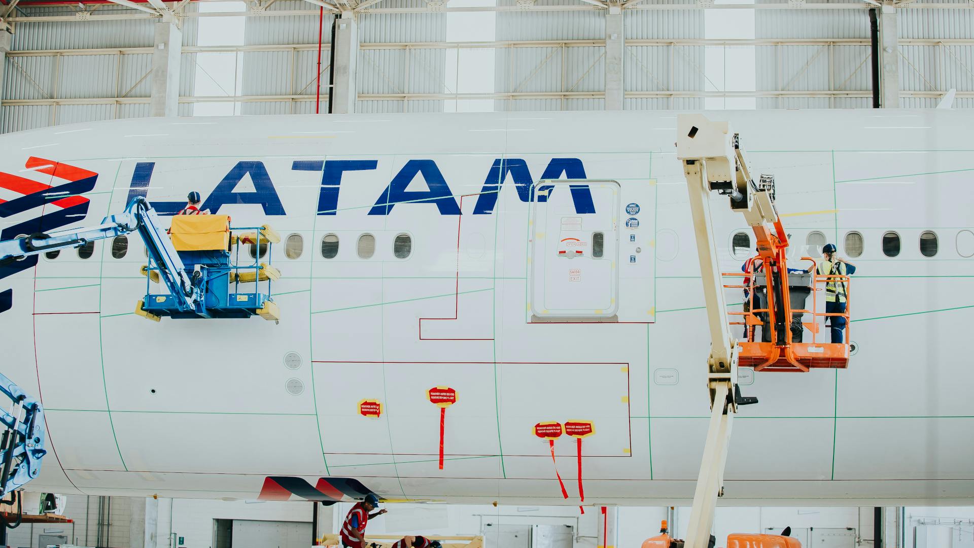 Technicians in lifted platforms applying a coating to a white airplane with text on it that reads: LATAM