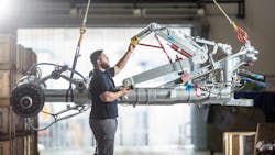 A technician working on a piece of airplane landing gear that's being suspended in the air by cables A technician working on a piece of airplane landing gear that's being suspended in the air by cables