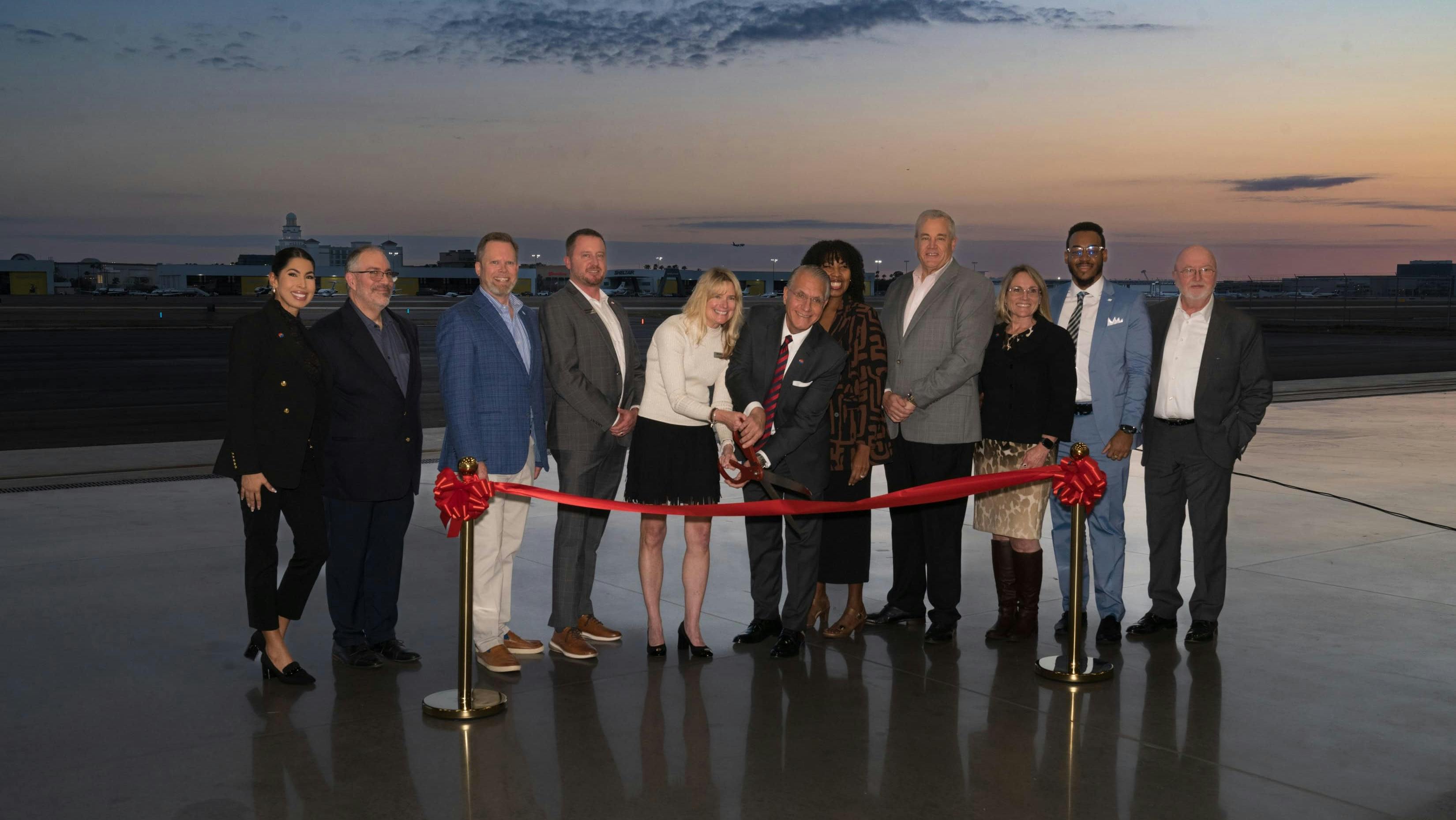 A group of people wearing business attire standing on a tarmac right after sunset during a ribbon cutting, with one woman holding large scissors over a red ribbon