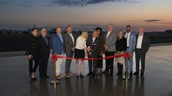 A group of people wearing business attire standing on a tarmac right after sunset during a ribbon cutting, with one woman holding large scissors over a red ribbon A group of people wearing business attire standing on a tarmac right after sunset during a ribbon cutting, with one woman holding large scissors over a red ribbon
