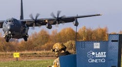 A black military airplane landing in front of a person wearing military fatigues and standing behind a sign that reads: LAST Armor QinetiQ A black military airplane landing in front of a person wearing military fatigues and standing behind a sign that reads: LAST Armor QinetiQ