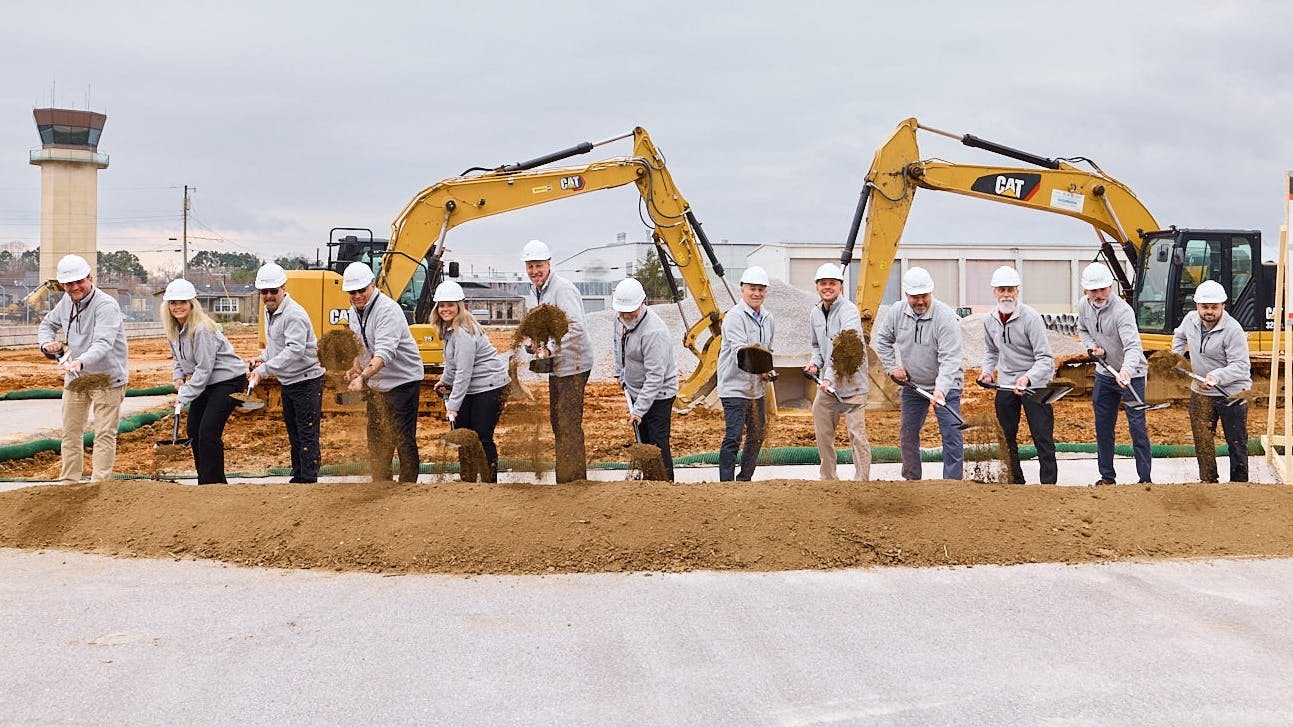 A group of professionals business attire and hard hats standing in a line holding shovels in front of a pile of soil, simulating a groundbreaking