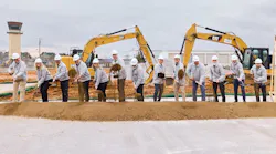 A group of professionals business attire and hard hats standing in a line holding shovels in front of a pile of soil, simulating a groundbreaking