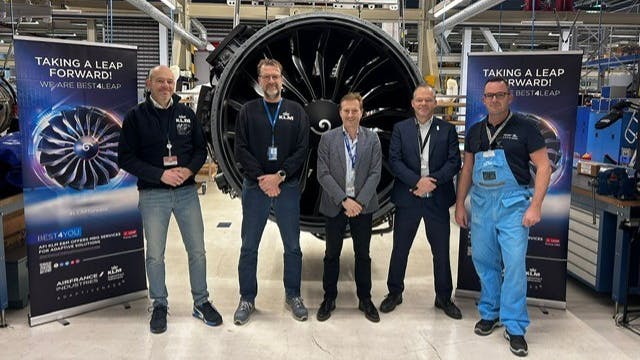 A group of men wearing ID badges standing in front of a detached aircraft engine next to signs that read: Take a leap forward