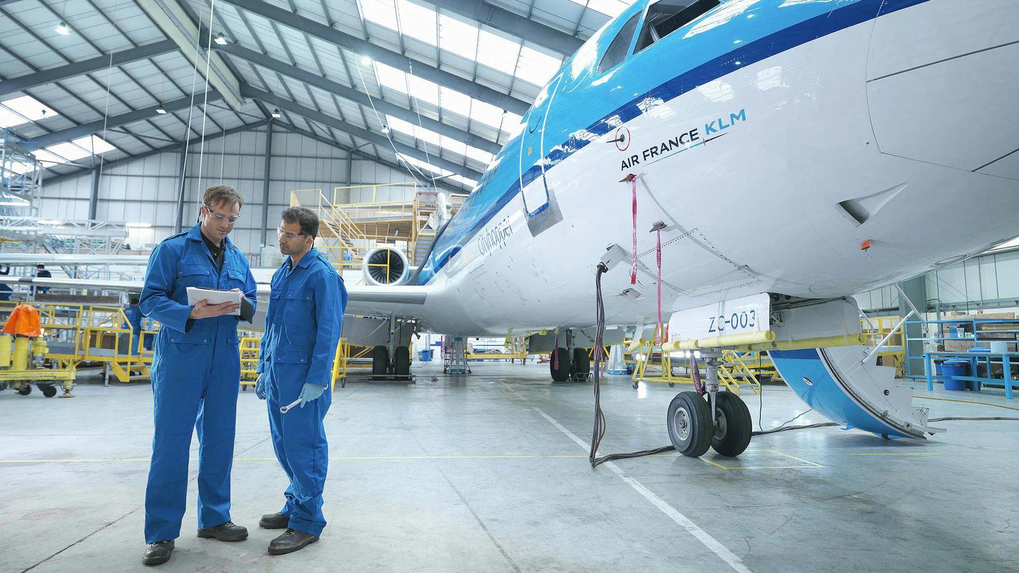 Two aircraft maintenance technicians wearing blue uniforms looking at a stack of papers while standing next to a blue and white airplane in a maintenance hangar