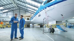 Two aircraft maintenance technicians wearing blue uniforms looking at a stack of papers while standing next to a blue and white airplane in a maintenance hangar Two aircraft maintenance technicians wearing blue uniforms looking at a stack of papers while standing next to a blue and white airplane in a maintenance hangar