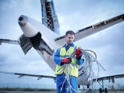 An aircraft technician wearing a blue uniform and yellow high-vis vest holding a bundle of cables while standing under an airplane wing An aircraft technician wearing a blue uniform and yellow high-vis vest holding a bundle of cables while standing under an airplane wing