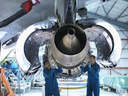 Two aircraft maintenance technicians wearing blue uniforms and protective goggles working inside of an open aircraft engine being held up on a stand Two aircraft maintenance technicians wearing blue uniforms and protective goggles working inside of an open aircraft engine being held up on a stand
