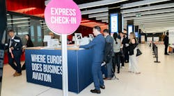 A line of professionals in business attire standing at a booth with a sign that says 'Express Check-In Where Europe Does Business Aviation' A line of professionals in business attire standing at a booth with a sign that says 'Express Check-In Where Europe Does Business Aviation'