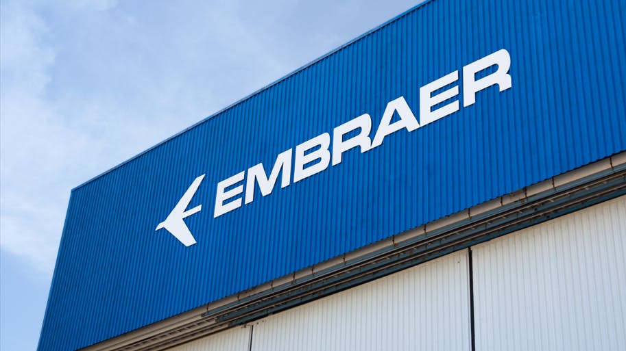 The side of a building with a beige wall and a dark blue sign with white lettering that reads: EMBRAER