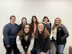 A group of women wearing casual clothing standing in front of a beige wall and smiling at the camera A group of women wearing casual clothing standing in front of a beige wall and smiling at the camera
