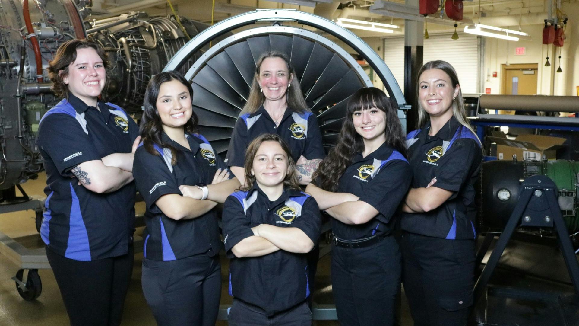 A group of female aircraft maintenance technicians wearing uniforms standing in front of an airplane engine and smiling at the camera