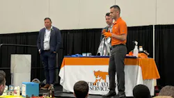 Three men standing on a stage with a table covered by a cloth that says: Foxtrot Aviation Services Three men standing on a stage with a table covered by a cloth that says: Foxtrot Aviation Services