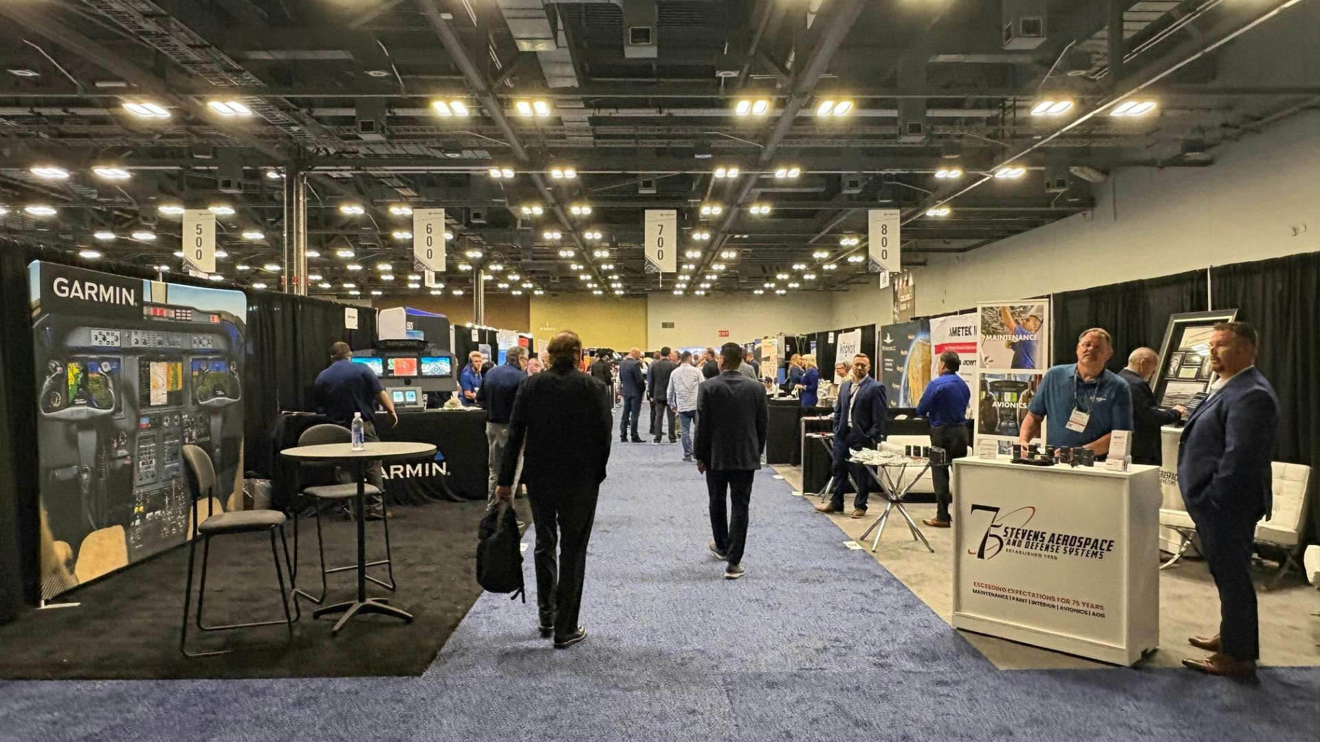 The floor of an exhibition hall with rows of tables and stalls, with professionals in business attire walking around the booths