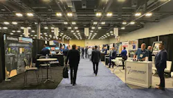 The floor of an exhibition hall with rows of tables and stalls, with professionals in business attire walking around the booths The floor of an exhibition hall with rows of tables and stalls, with professionals in business attire walking around the booths
