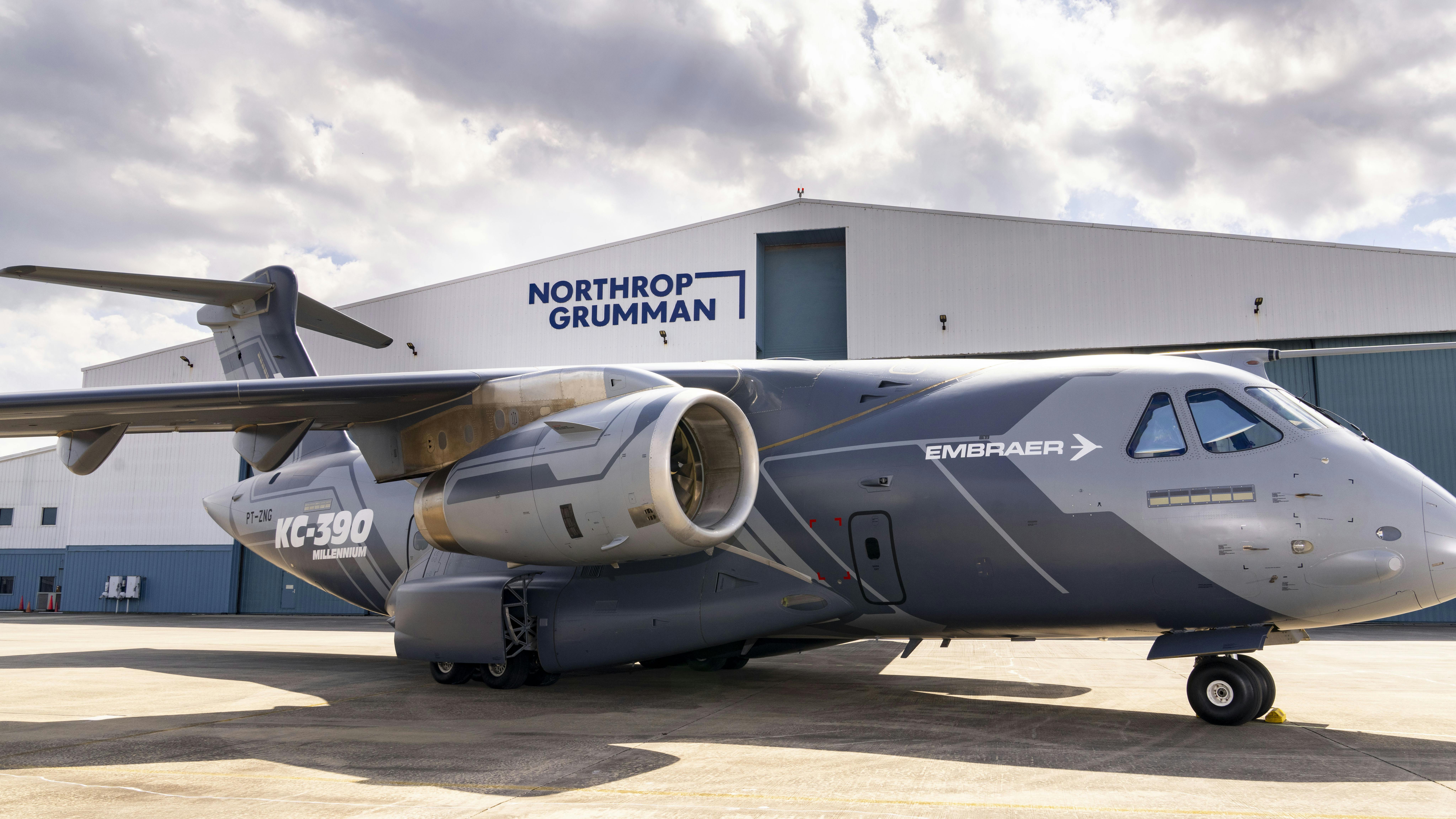 A gray airplane with text on it that reads 'EMRAER' and 'KC-390 Millennium' parked in front of a hangar with a sign on it that reads, 'Northrop Grumman'